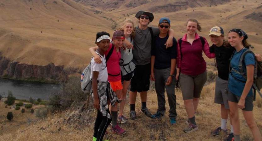 A group of students stand high above a river, wrapping their arms around each other and smile at the camera.